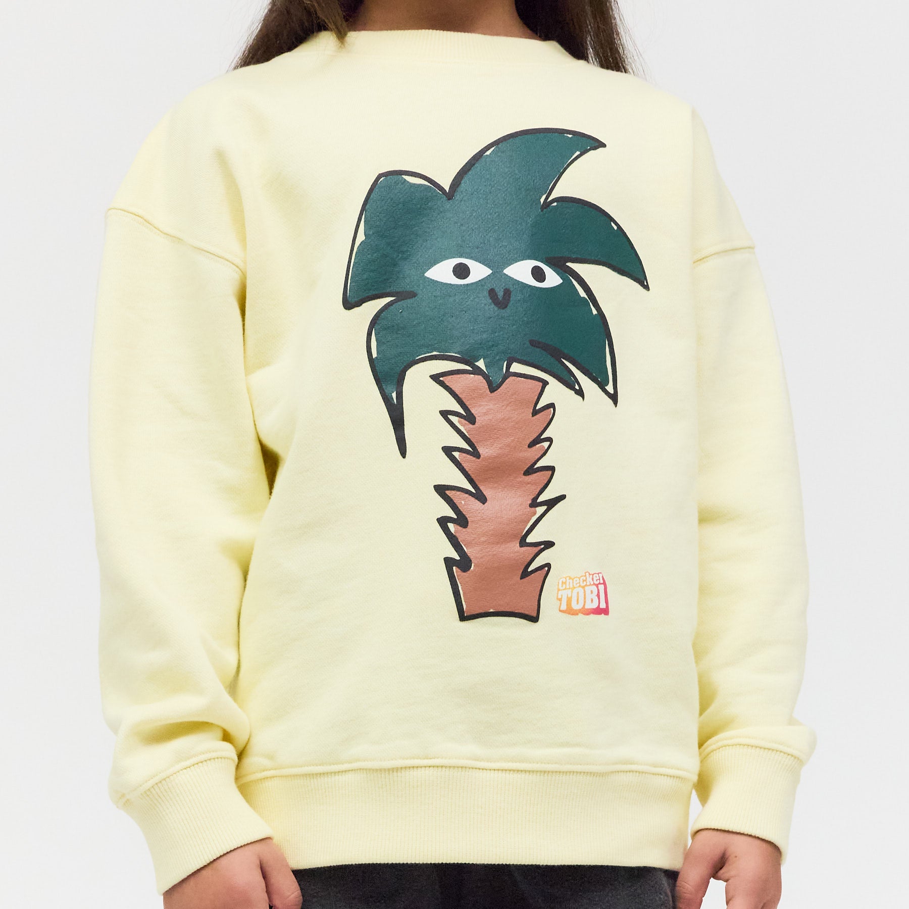 Nahaufnahme des Palmen und Checker Tobi Logo - Siebdrucks auf dem hellgelben Bio-Stoff – langlebige Farben und schadstofffreie Veredelung.