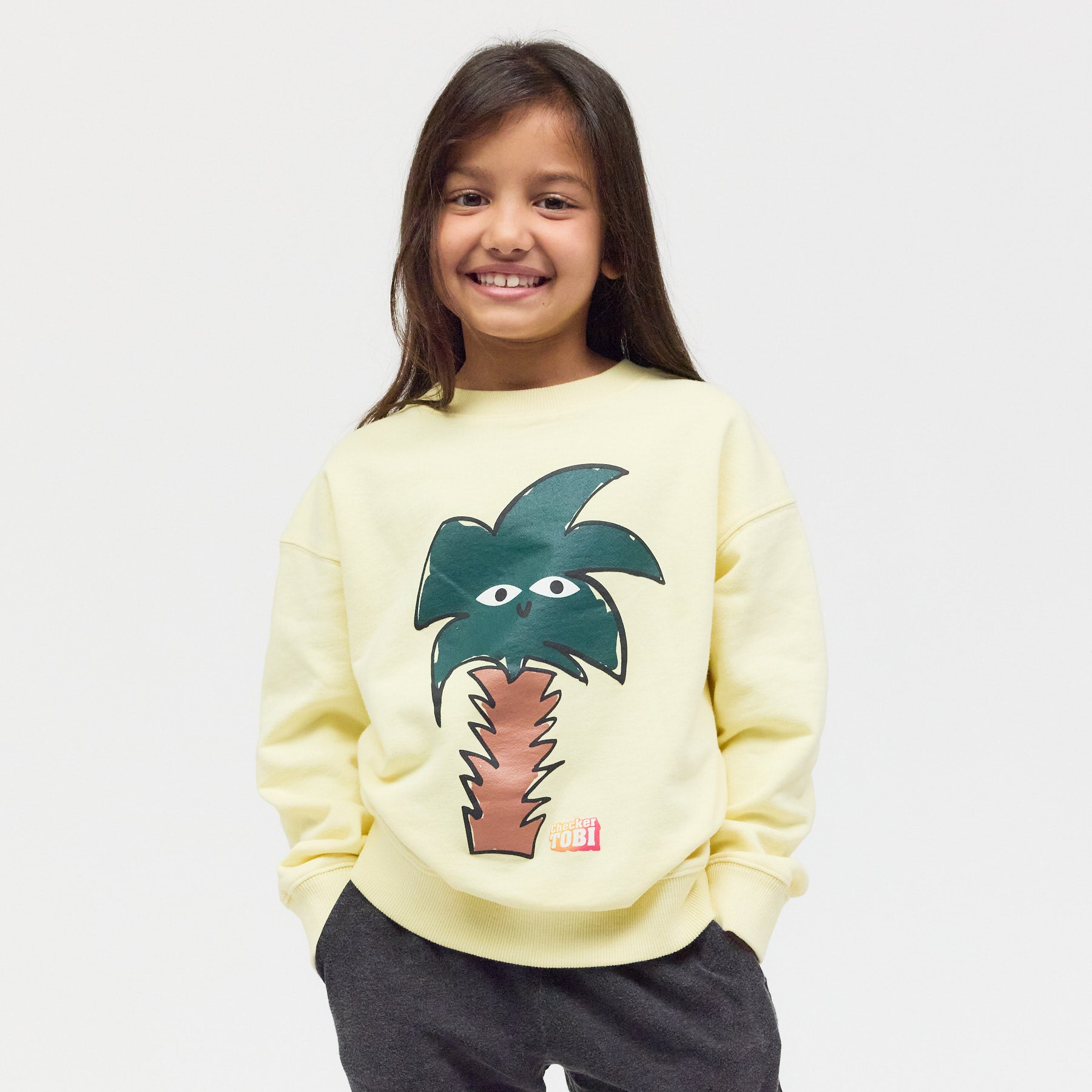 Kind trägt das hellgelbe Checker Tobi Sweatshirt mit Palmen-Grafik – gemütlicher Style aus atmungsaktiver Bio-Baumwolle für kleine Entdecker.