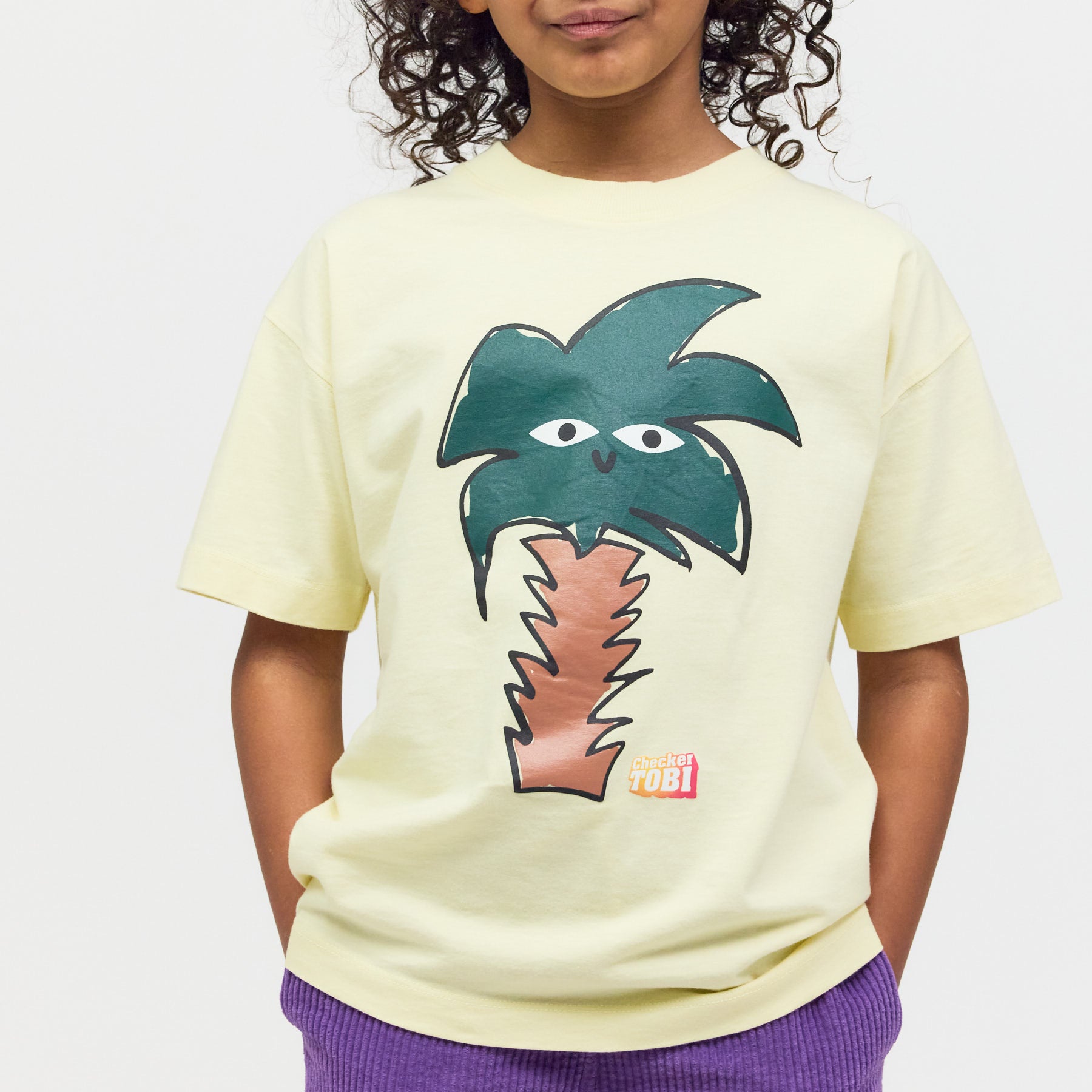 Kind trägt das gelbe Checker Tobi T-Shirt mit Palmen-Print – weicher Bio-Stoff und schadstofffreier Druck für maximalen Tragekomfort.